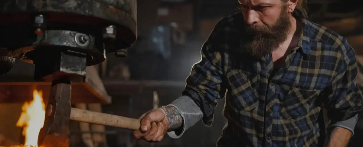 A bearded man in a blue and black plaid shirt uses a wooden handle to turn a large forge or anvil in a dark workshop, with a bright flame visible on the left.